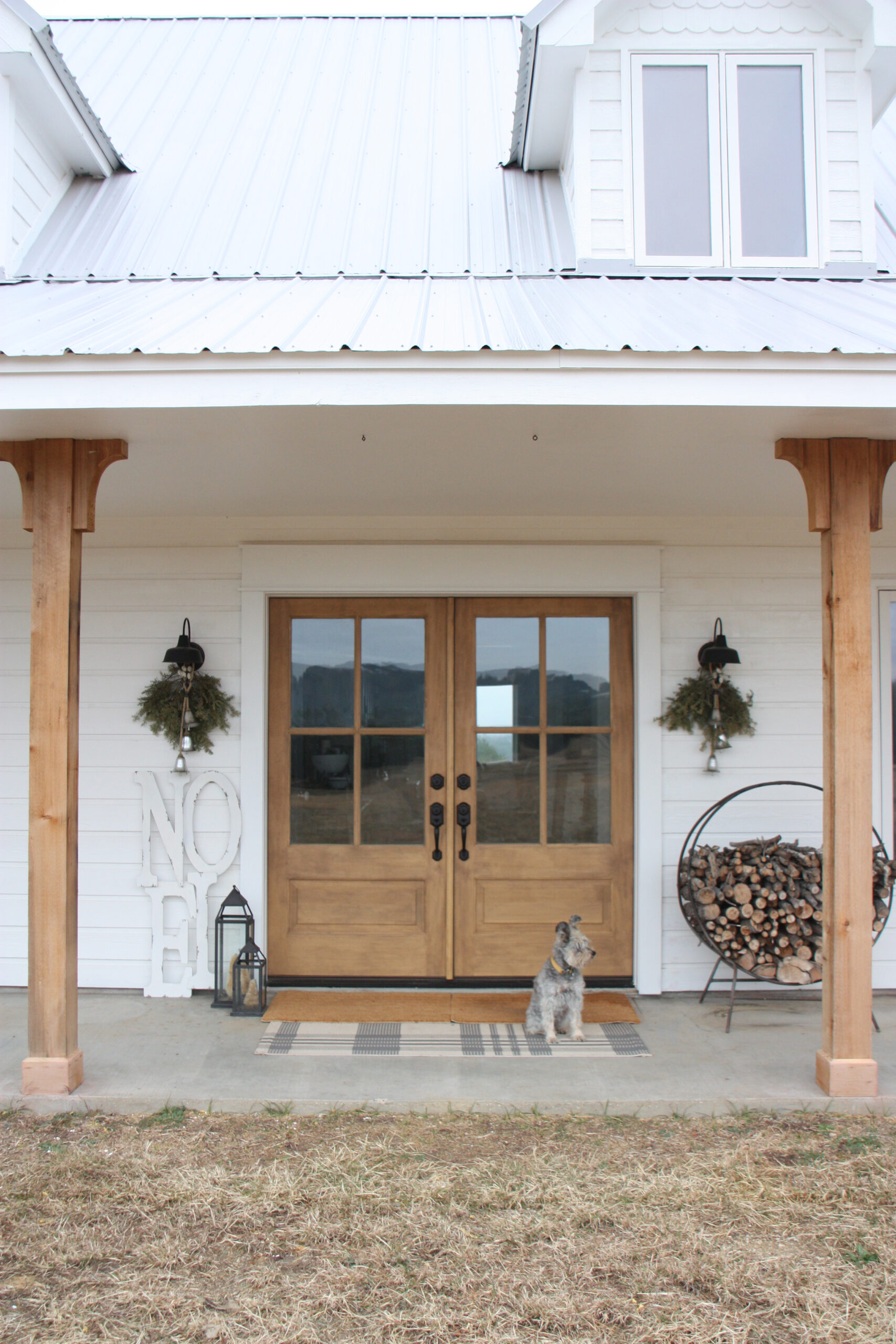 White farmhouse porch with light wood double entry doors, wreaths, stacked firewood and a dog