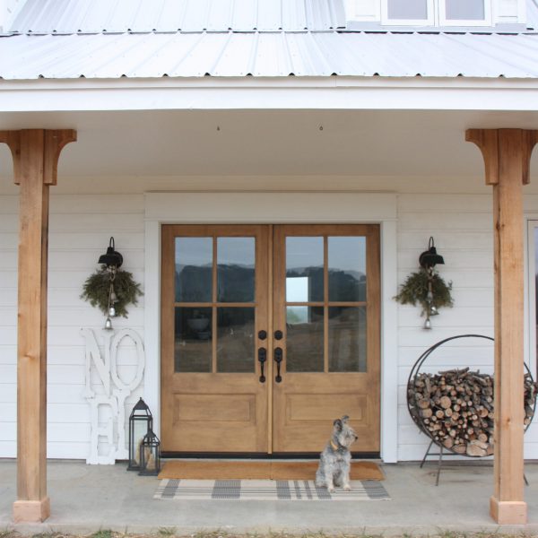 White farmhouse porch with light wood double entry doors, wreaths, stacked firewood and a dog